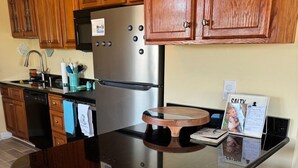 A kitchen with wooden cabinets, a stainless steel refrigerator, a black countertop, and a small island with a woven chair.