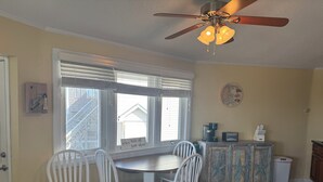 A dining area with a round table, four white chairs, a ceiling fan, and a window with blinds.