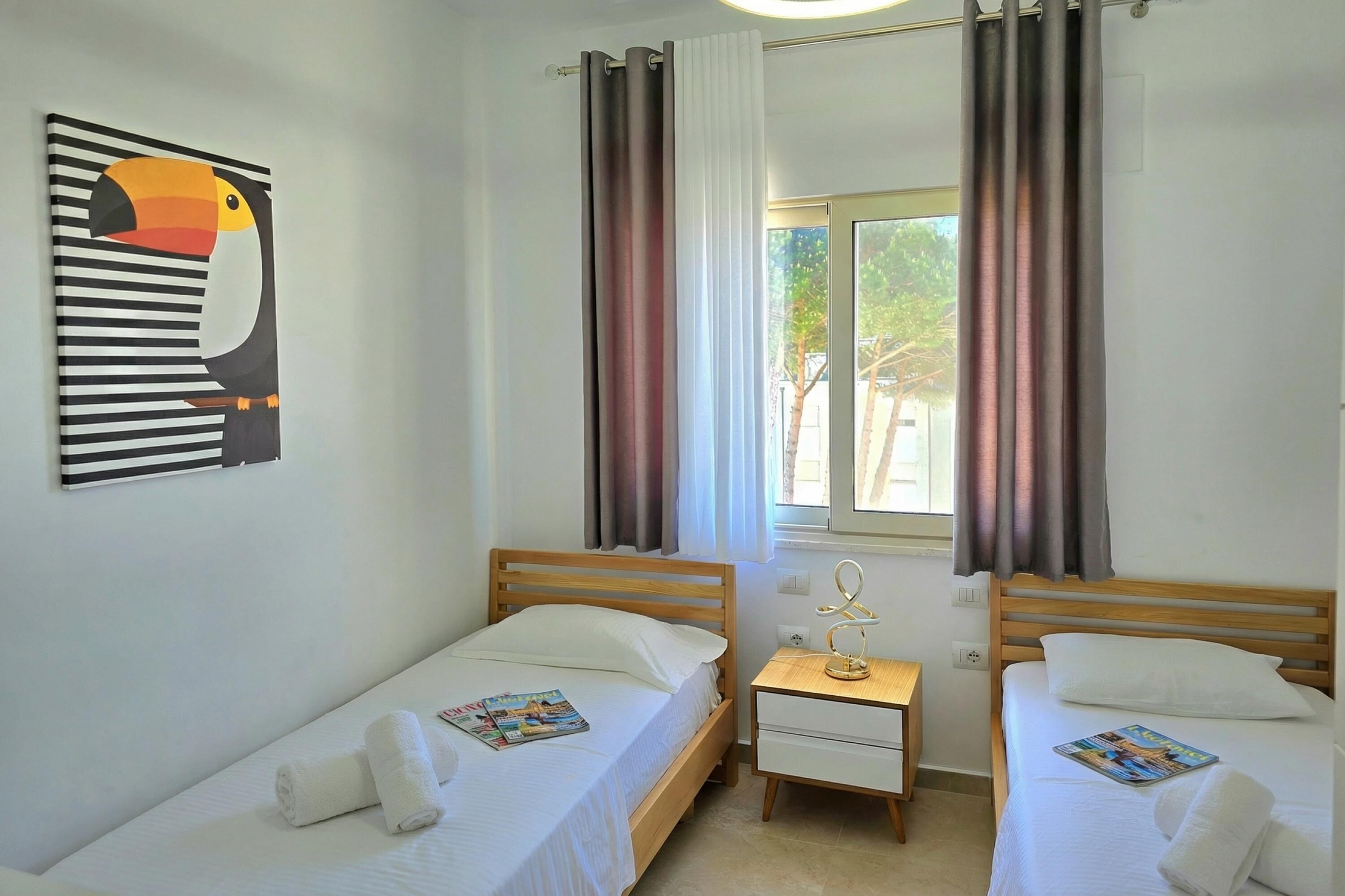 A room with two single beds, a wooden nightstand, a wall-mounted artwork, and a window with curtains.