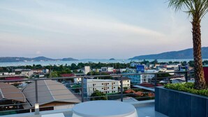 A rooftop terrace with a round table and white chairs, overlooking a coastal town with buildings and boats. - Garden View Apt- 1BR T1-103 (Chalong)