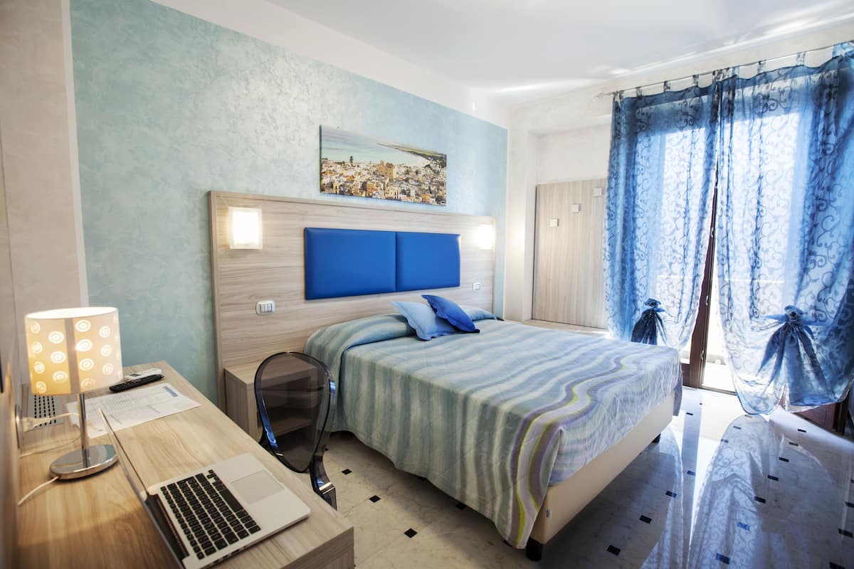 A hotel room with a bed, a desk with a laptop, a chair, and a lamp.
