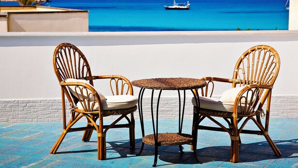 A terrace with two wicker chairs and a small table, overlooking a clear blue sea with a sailboat in the distance. - Hotel Piccolo Mondo (San Vito Lo Capo)