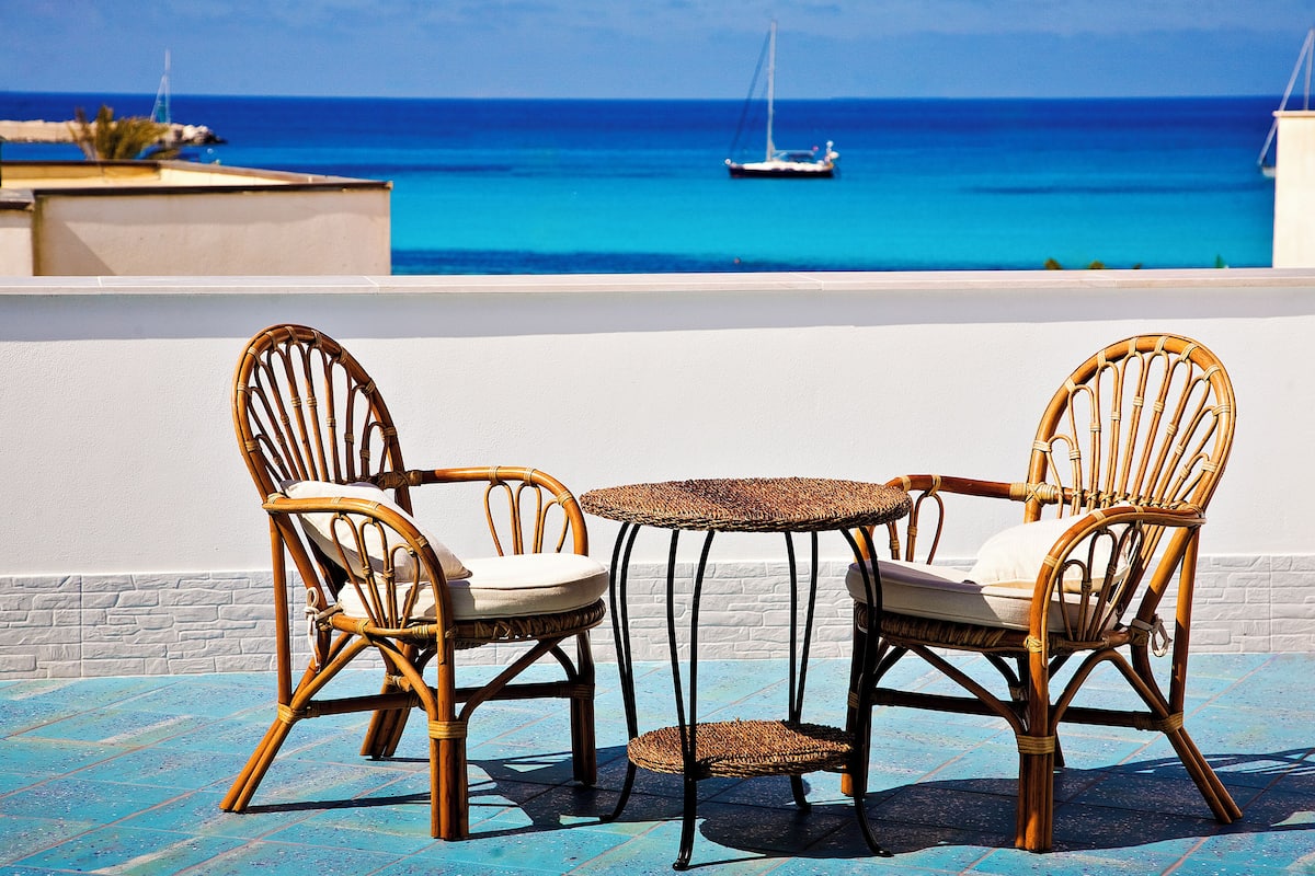 A terrace with two wicker chairs and a small table, overlooking a clear blue sea with a sailboat in the distance.