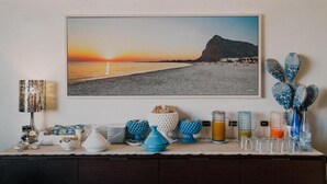 A wall-mounted photograph of a beach at sunset, a decorative table with various items, and a patterned lamp. - Hotel Piccolo Mondo (San Vito Lo Capo)