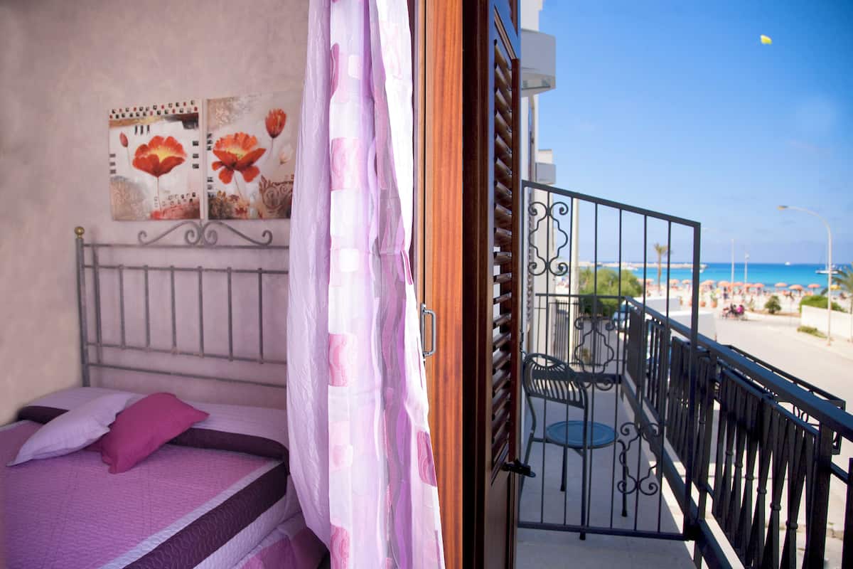 A balcony with a metal railing, a bed with pink bedding, and a view of the beach.
