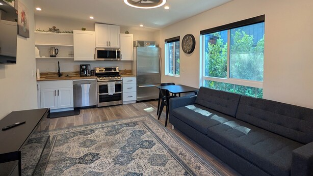 A modern kitchen with white cabinets, a stainless steel refrigerator, and a dining area with a dark sofa and rug. - Guesthouse in Anaheim - Monthly Rental (Anaheim)