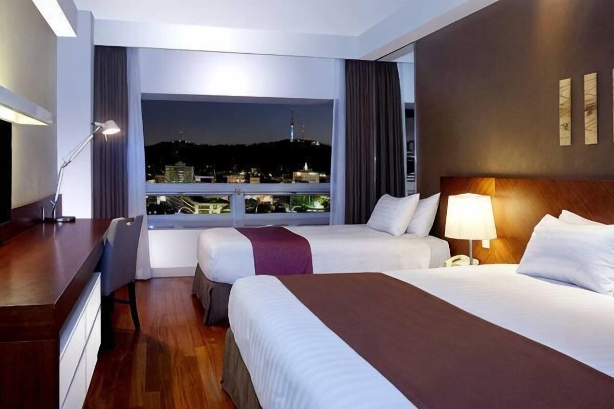 A hotel room with two beds, a desk, a chair, a lamp, and a view of a cityscape at night.