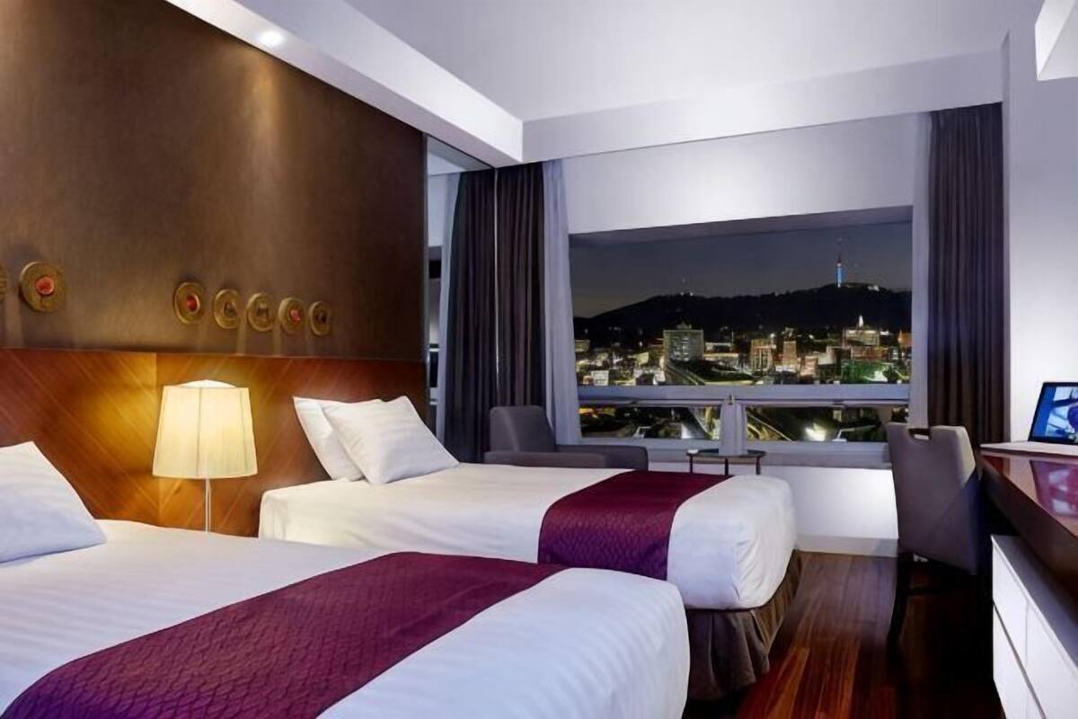 A hotel room with two beds, a desk, a chair, a lamp, and a large window with a city view.