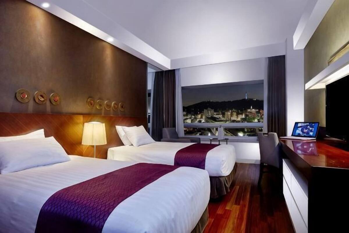 A hotel room with two beds, a desk, a TV, and a city view.