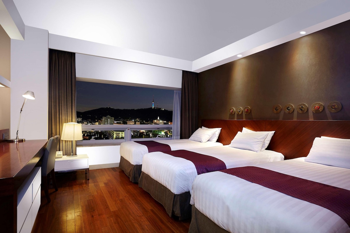 A hotel room with three beds, a desk, and a view of a cityscape at night.