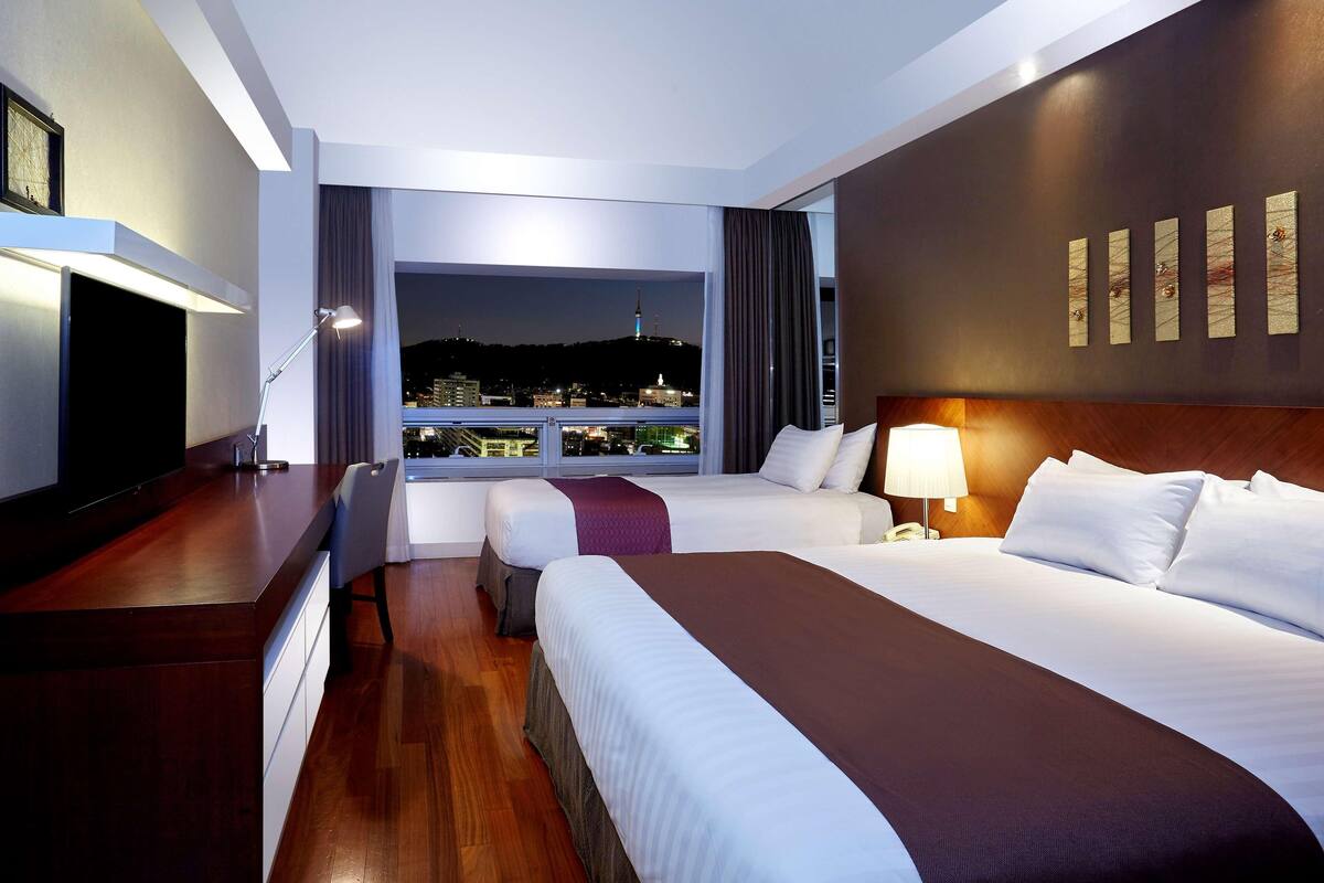 A hotel room with a large bed, a smaller bed, a desk with a TV, a window with a city view, and a wall with decorative panels.