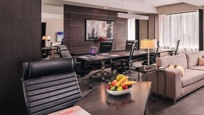 A modern hotel room with a dark wood desk, a leather chair, a sofa, and a dining table with chairs. There is a bowl of fruit on the desk and a lamp on the table.
