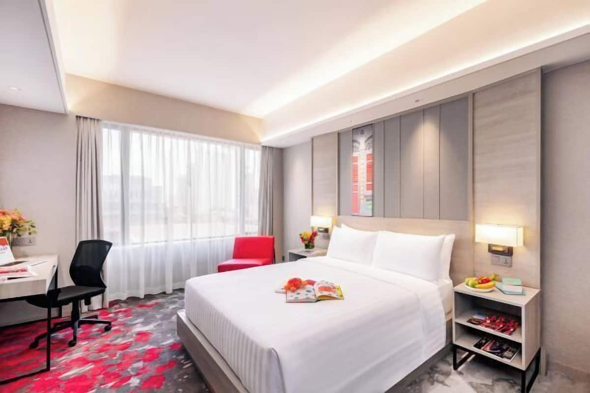 A hotel room with a large bed, a desk with a chair, a red chair, a nightstand with a lamp, and a window with curtains.