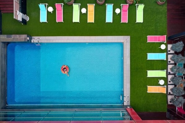A swimming pool with a floating pool toy, a green artificial grass wall, and a row of colorful towels.