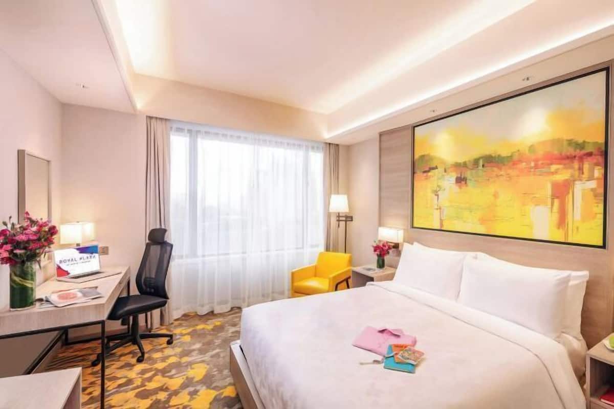 A hotel room with a bed, a desk with a chair, a small table with a lamp, and a large painting on the wall.