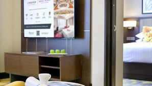 A hotel room with a television displaying a room image, a wooden cabinet with a tray of apples, and a bed with a floral bedspread.
