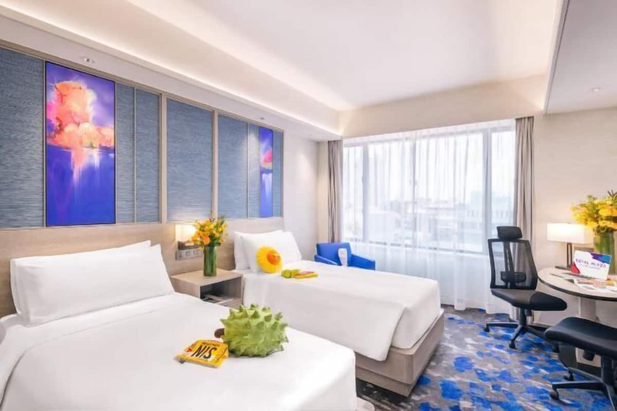 A hotel room with two beds, a desk, a chair, a large window, and a wall with artwork.