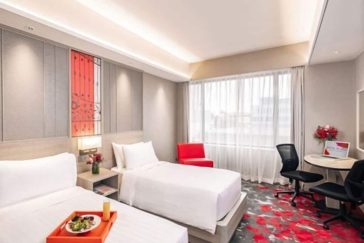 A hotel room with two beds, a desk, a chair, a small table with a laptop, and a window with curtains.