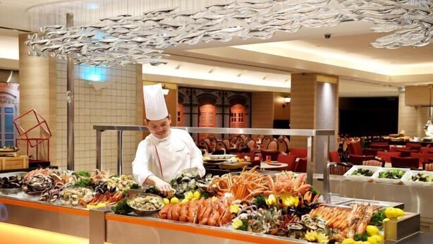 A seafood buffet with a variety of shellfish and seafood dishes, a chef in uniform, and a modern interior with red chairs and a tiled wall.