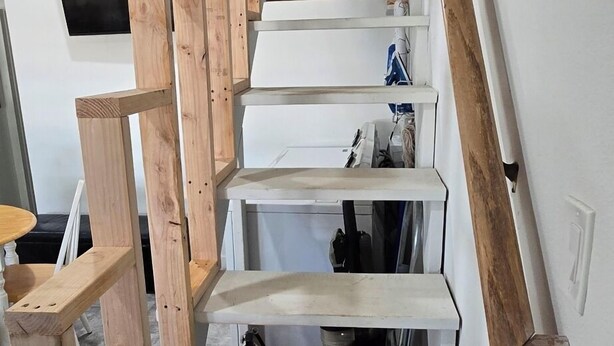 Eine Holztreppe mit weißen Stufen und einem Geländer, die zu einem Zwischenpodest mit einem Feuerlöscher und einem an der Wand befestigten Fernseher führt.