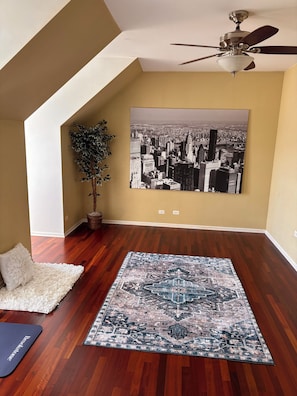 A room with a cityscape painting, a ceiling fan, a potted plant, a white fluffy rug, and a patterned rug on a wooden floor.