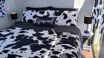 Una cama con colcha y almohadas estampadas con vaca en blanco y negro, un letrero que dice "Unwind" y una mesita de noche con una lámpara.