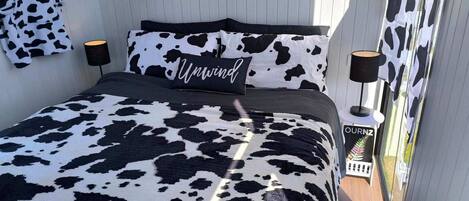 A bed with a black and white cow print bedspread and pillows, a sign reading "Unwind", and a small side table with a lamp.