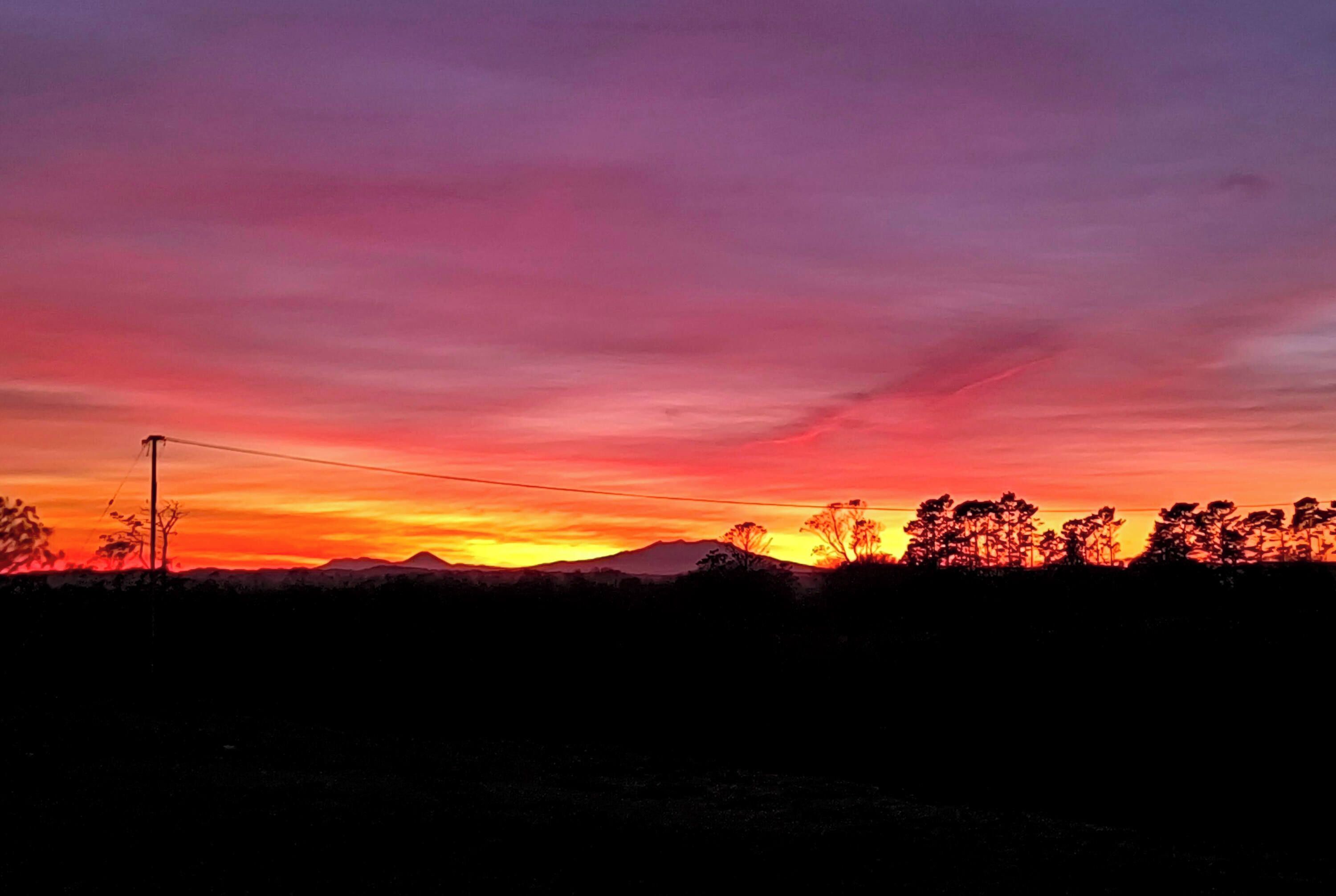 A sunset over a landscape with silhouettes of trees and distant mountains.