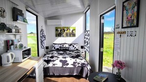 A bedroom with a cow-print bedspread, a kettle, a bookshelf, and a window with a view of the outdoors.
