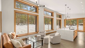 Een ruime woonkamer met grote ramen, een centraal keukenblok met spoelbak, en een moderne kroonluchter.