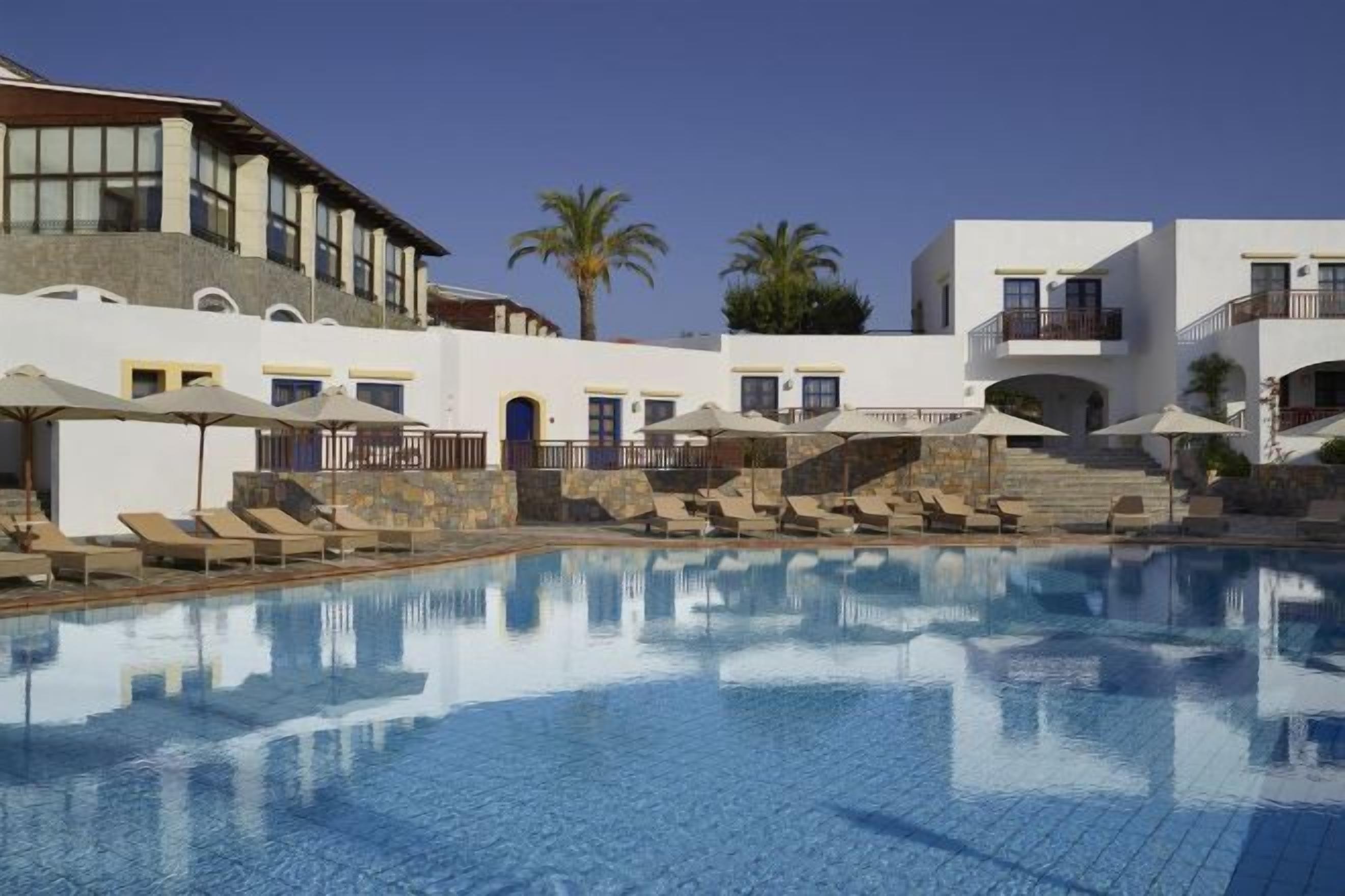 A swimming pool with lounge chairs and umbrellas, surrounded by buildings with balconies and a clear sky.
