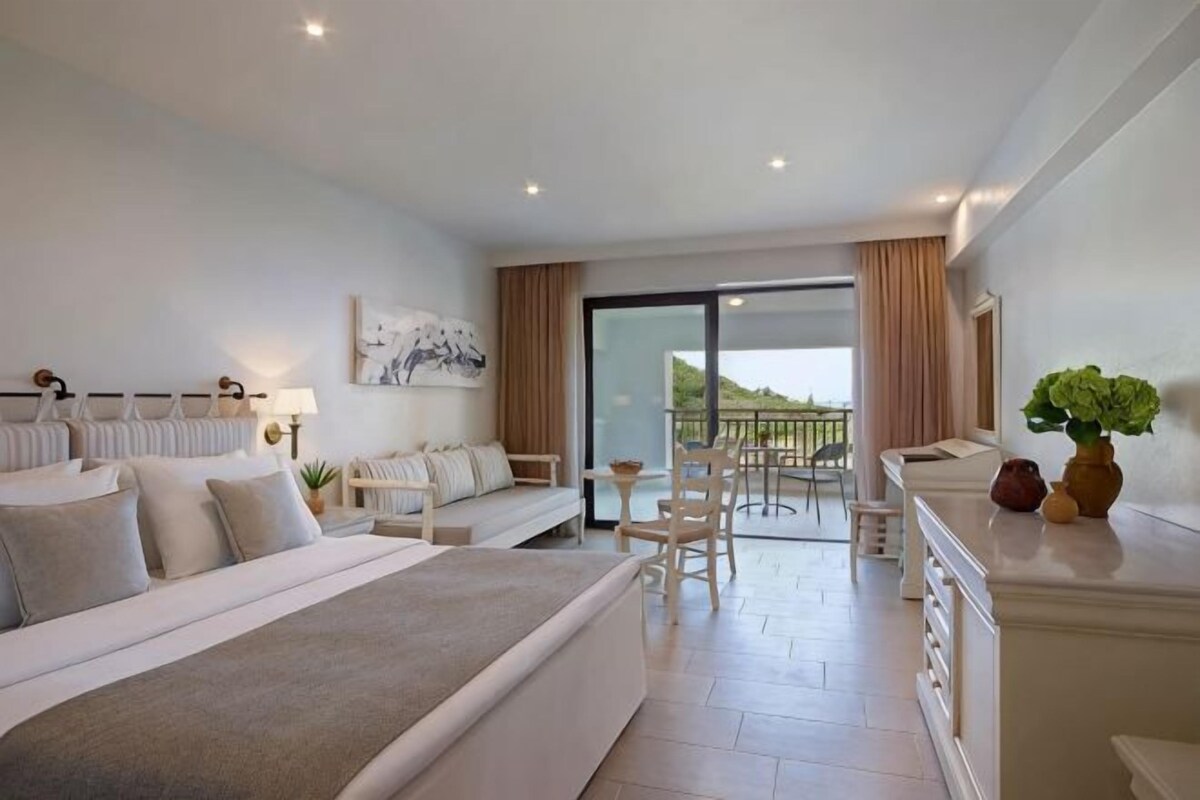 A hotel room with a bed, seating area, balcony, and a view of the outdoors.