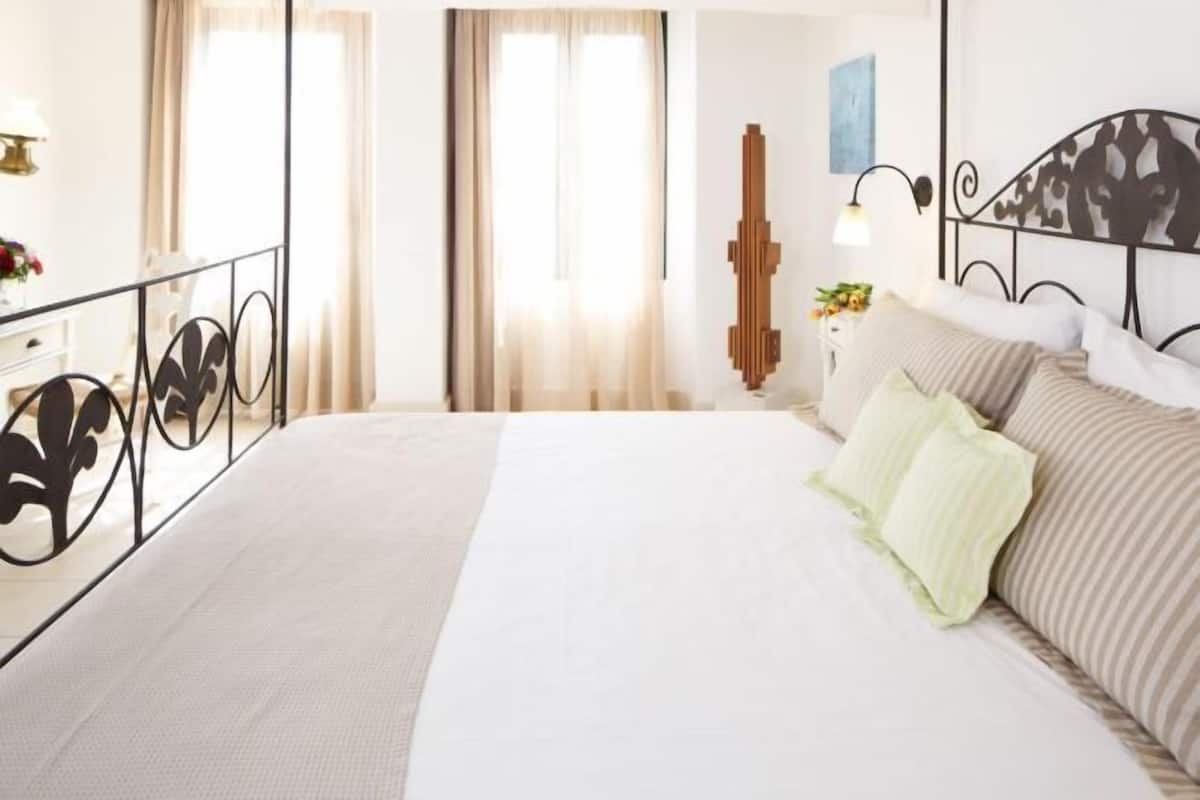Une chambre à coucher avec un lit, des rideaux, une table de chevet, une lampe et une œuvre d’art accrochée au mur.