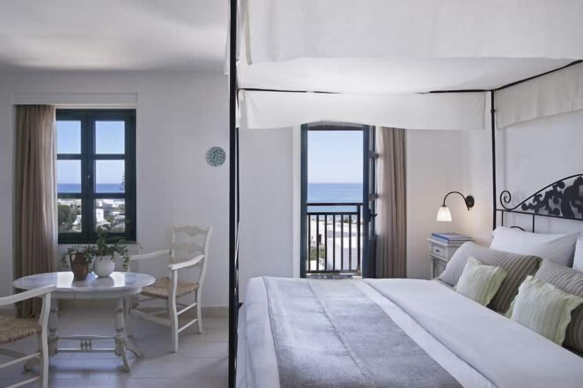 Une chambre à coucher avec un lit, une petite table et des chaises, une fenêtre donnant sur la mer et une porte-fenêtre donnant sur un balcon.