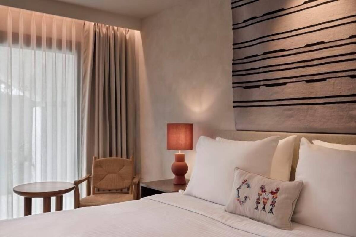 Une chambre d’hôtel avec un lit, une chaise, une petite table, une lampe et une grande fenêtre avec des rideaux.