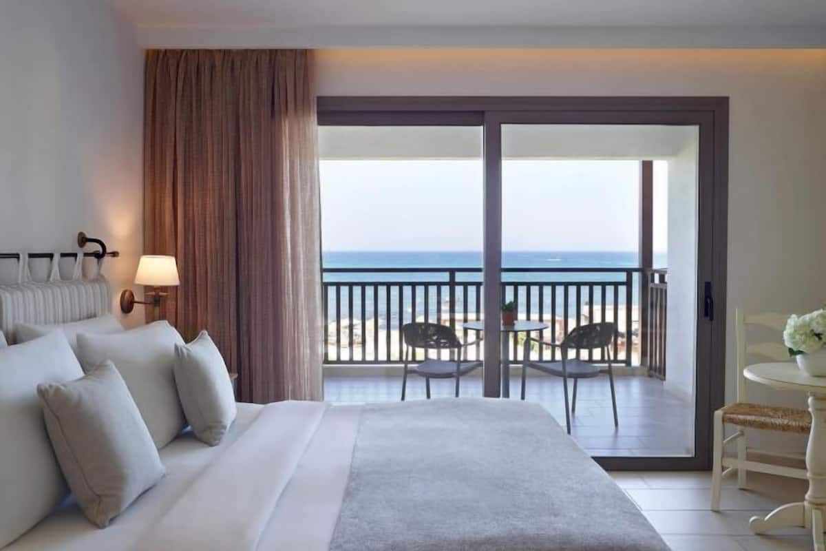 Une chambre d’hôtel avec un lit, une table de chevet, une lampe, une vue sur la mer et un balcon équipé de chaises et d’une table.