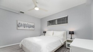 Une chambre à coucher avec un lit, deux tables de chevet avec des lampes, un ventilateur de plafond, une fenêtre avec des stores et un tableau au mur.