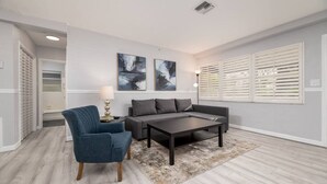 Un salon moderne avec un canapé gris, un fauteuil bleu et une table basse. Deux tableaux abstraits ornent le mur, et une fenêtre est équipée de stores.