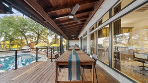 Een terras buiten met een houten tafel en stoelen, een zwembad en grote ramen met uitzicht op bomen en een gebouw.