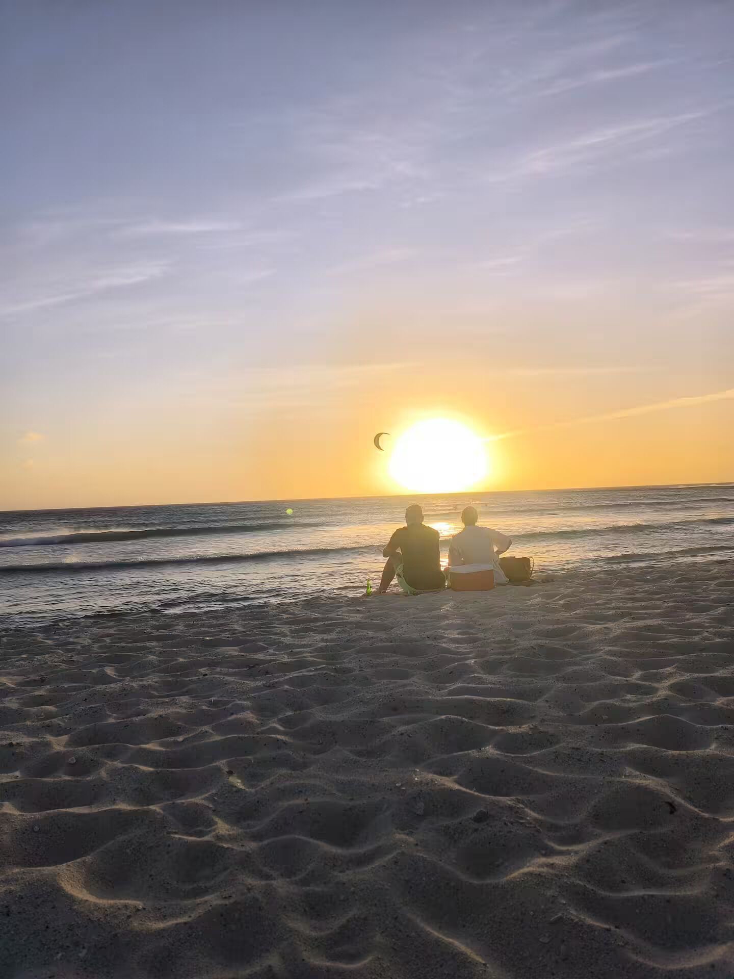 Two people sitting on the beach facing the sunset.
