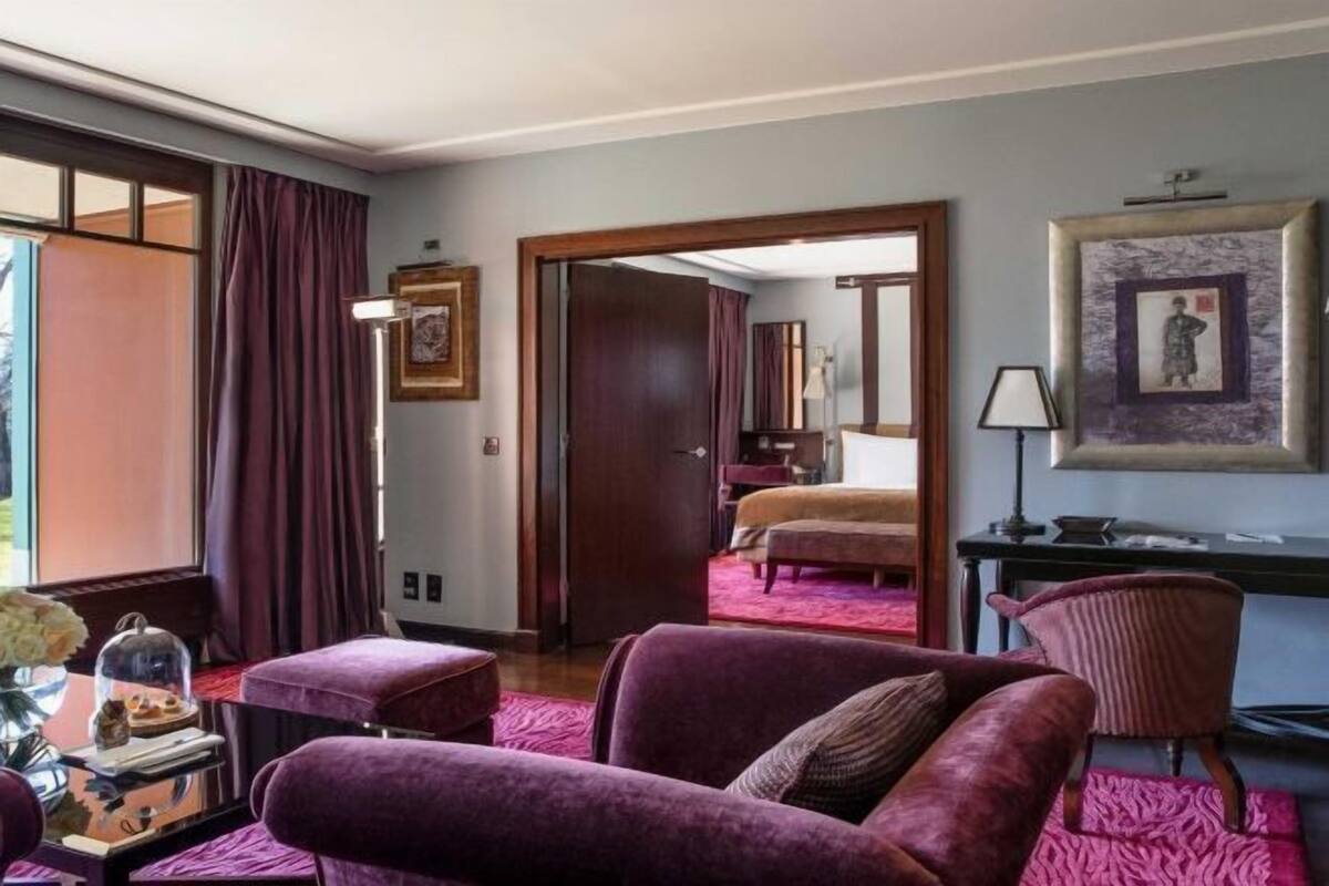 Una habitación de hotel con un sofá morado, un escritorio, una silla, una cama y un amplio ventanal con cortinas.