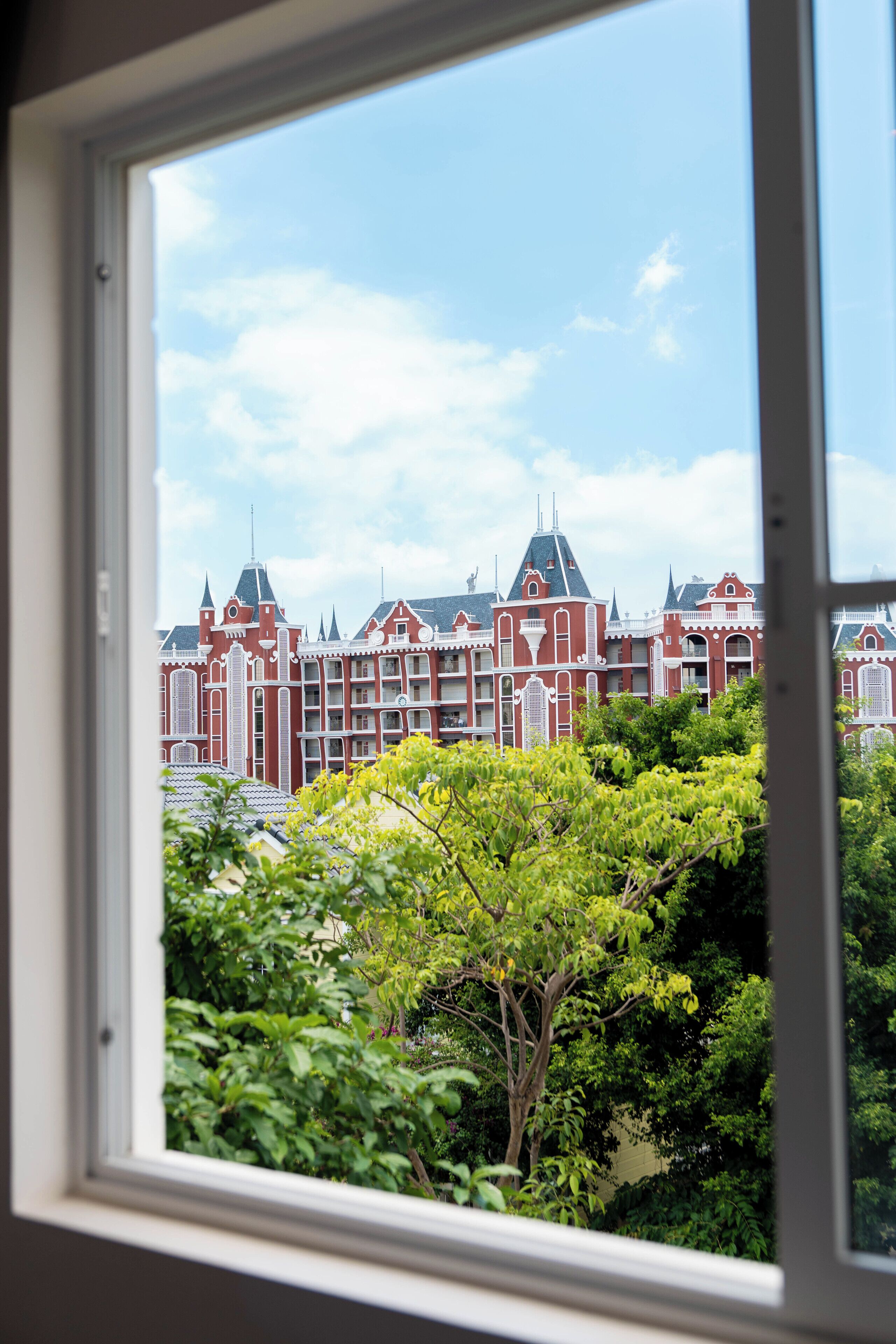 Vista a través de una ventana que muestra un edificio con múltiples detalles arquitectónicos en rojo y blanco, incluyendo torres y agujas, y abundante follaje verde en primer plano.