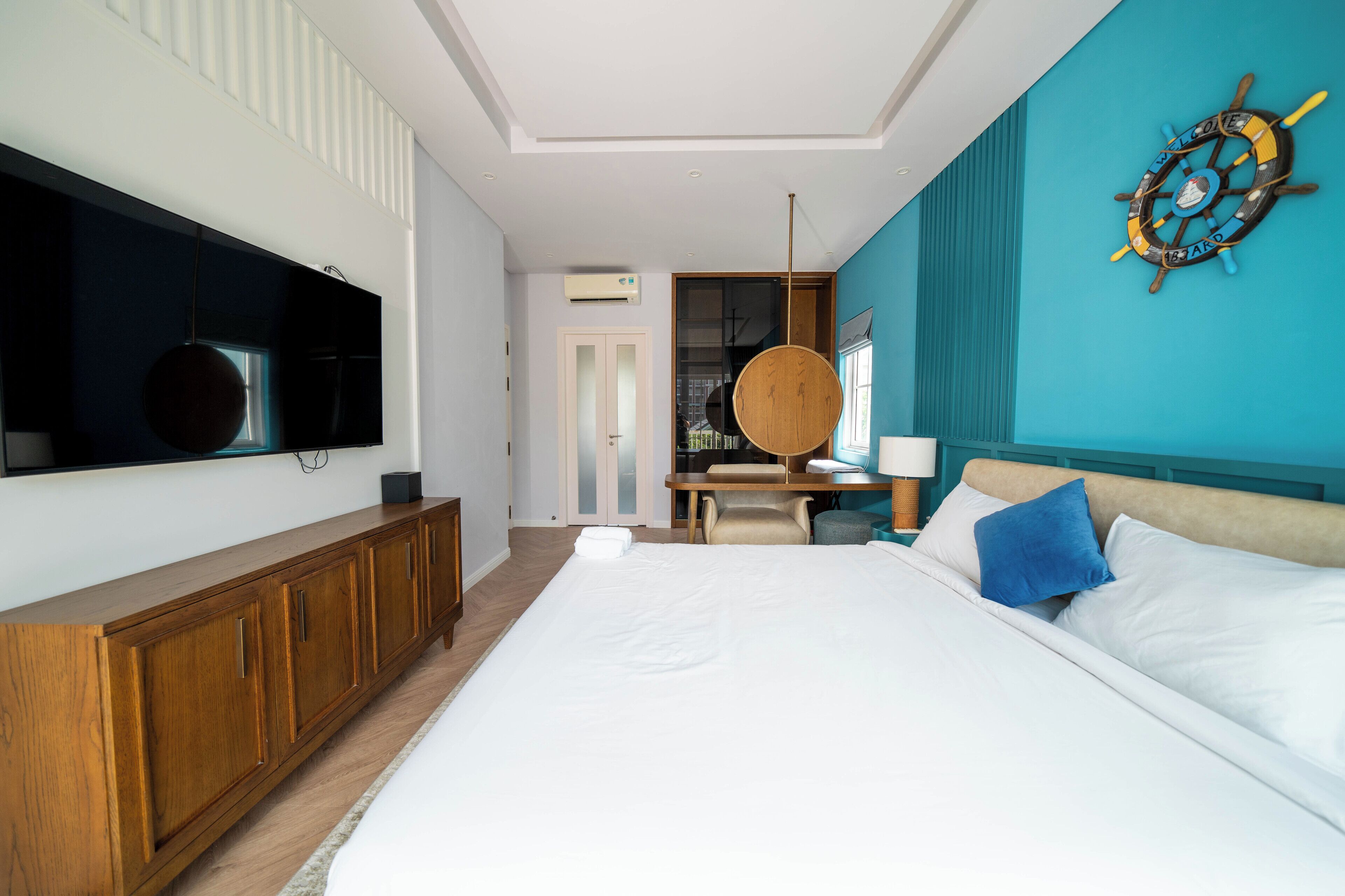 A modern hotel room with a large bed, a wooden wardrobe, a desk with a chair, a television mounted on the wall, and a ship's wheel decoration.