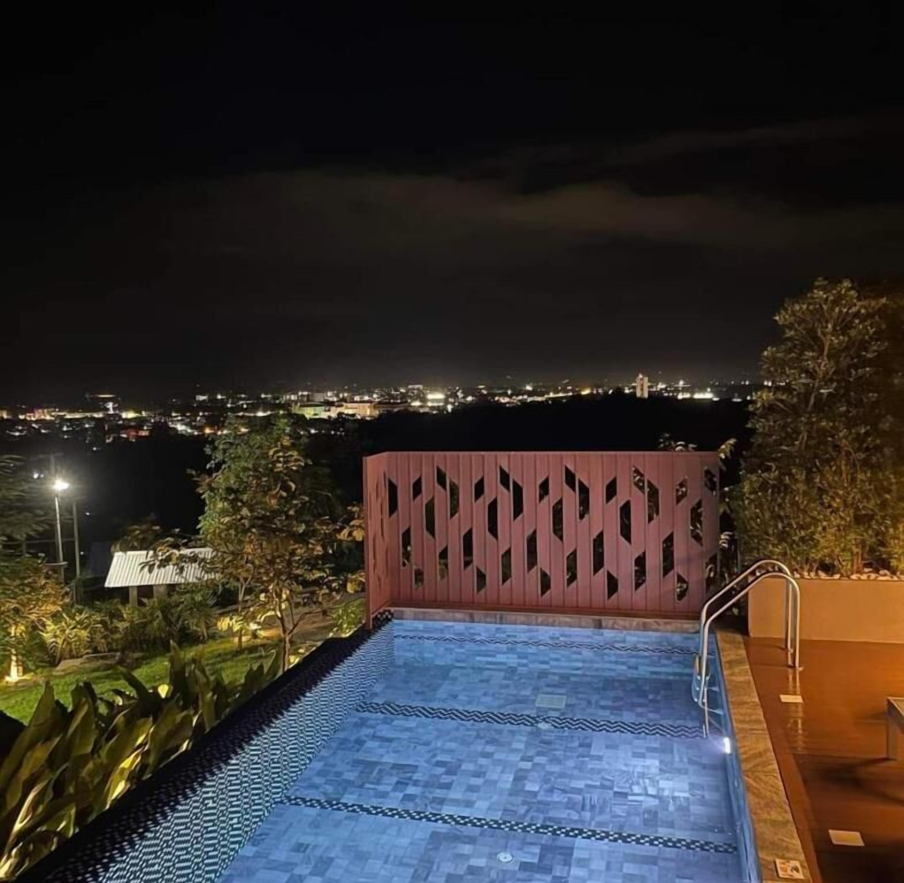 A swimming pool with a patterned floor, surrounded by greenery and a view of a cityscape at night.