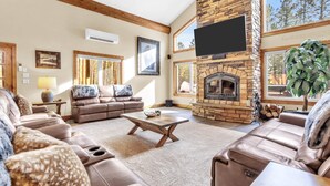 A spacious living room with leather sofas, a stone fireplace, and large windows offering a view of trees.