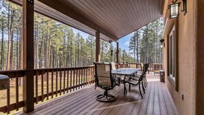 A covered outdoor deck with a table and chairs, surrounded by a wooden railing, overlooking a forested area.