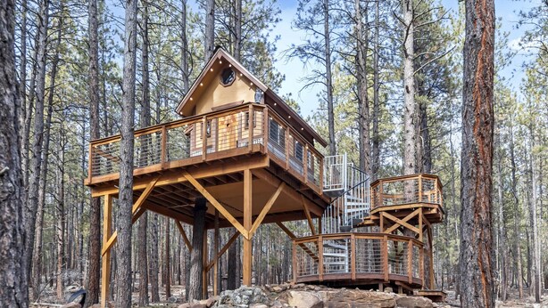 A wooden treehouse with multiple levels and a small cabin-like structure, surrounded by a forest.