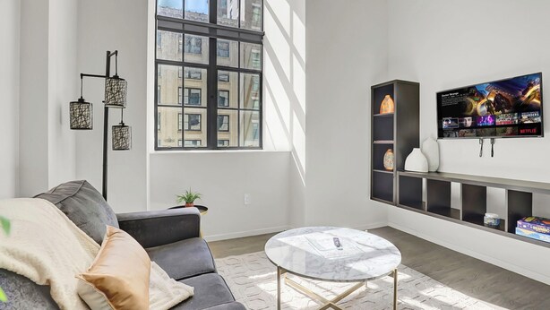 A modern living room with a grey sofa, a round coffee table, a large window with city views, a wall-mounted TV, and a black shelving unit. - Chic 1-bed loft in Playhouse square, near Stadiums, Theater, Dining, Pets OK (Cleveland)
