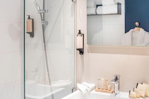 Une salle de bain moderne avec une douche à l’italienne en verre, un lavabo blanc agrémenté d’articles de toilette et un miroir qui reflète un lit soigneusement fait avec du linge de lit blanc.