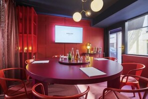 Un salón de conferencias con una mesa redonda, sillas rojas, una pantalla de proyector y vistas al exterior.